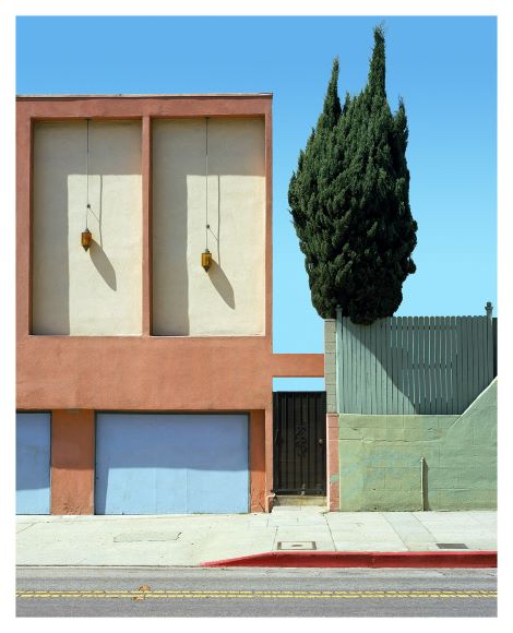 A minimalist building facade with two light fixtures, adjacent to a tall, narrow tree and a green wooden fence under a clear blue sky.