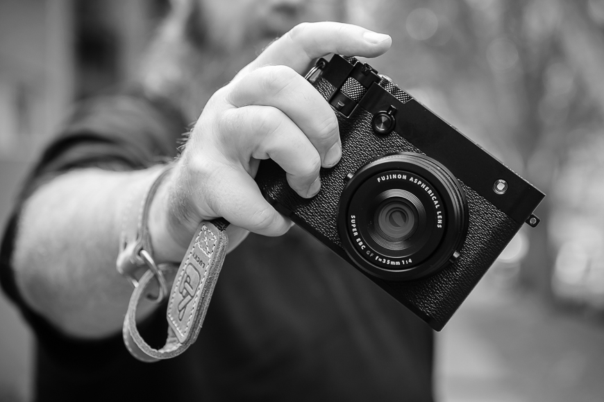 Person holding a Fujifilm camera towards the viewer in an outdoor setting.