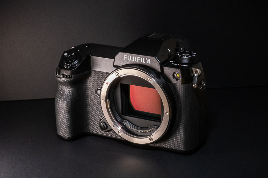 A black Fujifilm camera without a lens attached is displayed on a dark surface against a black background.