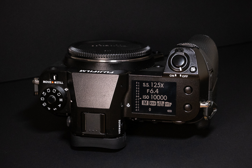 Top view of a Fujifilm digital camera showing its control dials and LCD screen displaying settings like shutter speed, aperture, and ISO.
