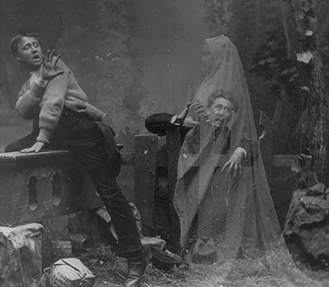 Two men appear startled by a ghostly figure draped in a translucent sheet, set outdoors with trees and a stone bench in the background.