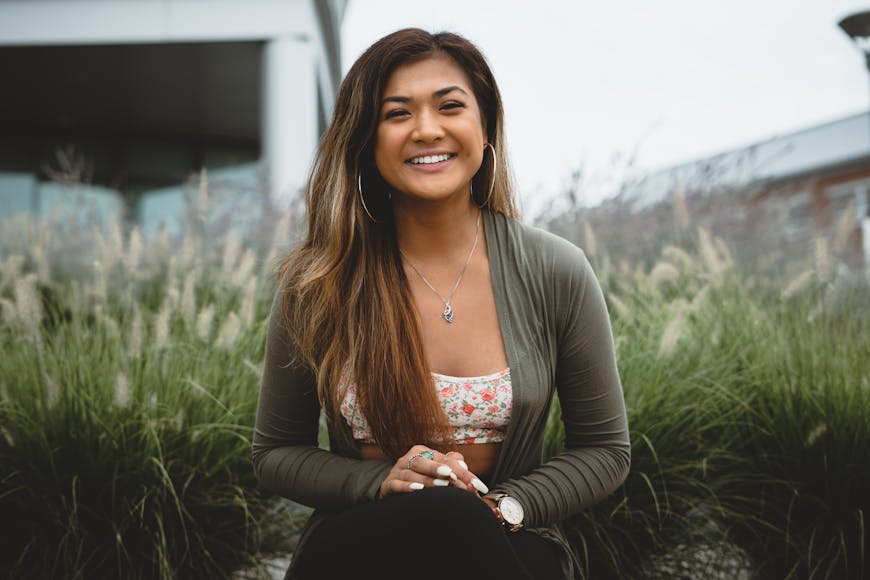 A woman with long brown hair sits outdoors in front of tall grass, smiling at the camera. She wears a green cardigan over a floral top and has hoop earrings.
