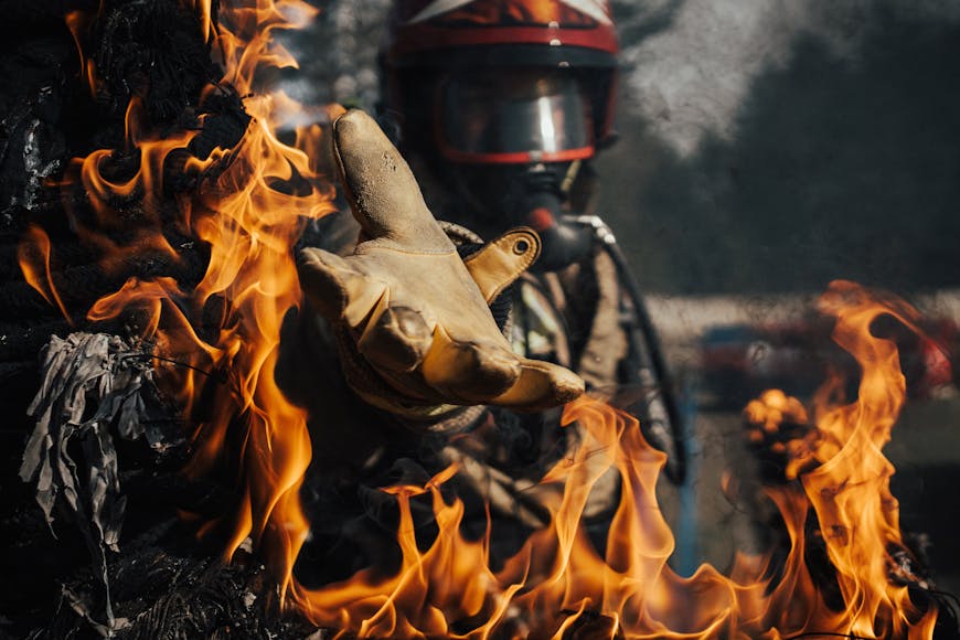 A firefighter in protective gear reaches through flames with a gloved hand during a fire emergency.