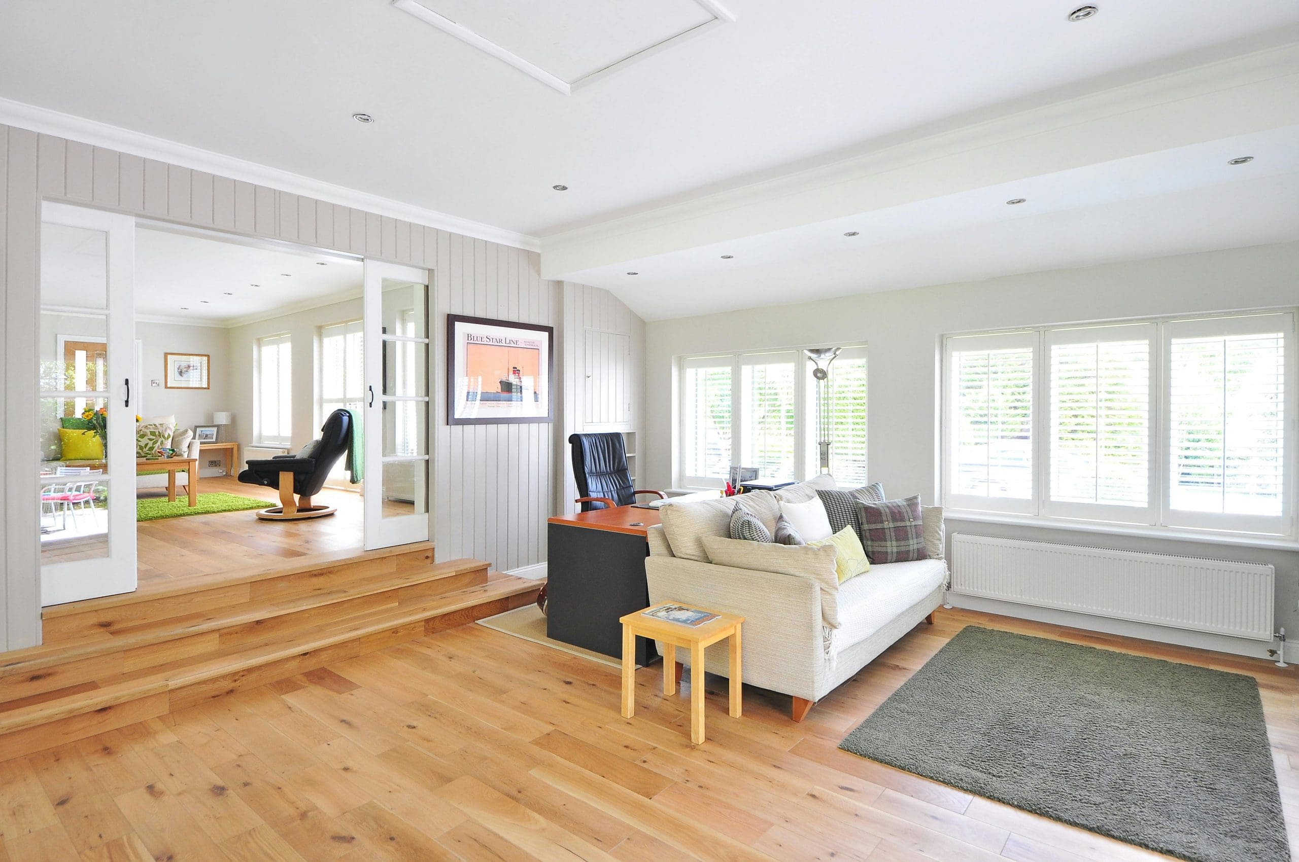 A bright, open living area with wooden floors, white walls, a beige sofa, a green rug, and a black office chair next to a desk. Sunlight streams through the large windows with white frames.