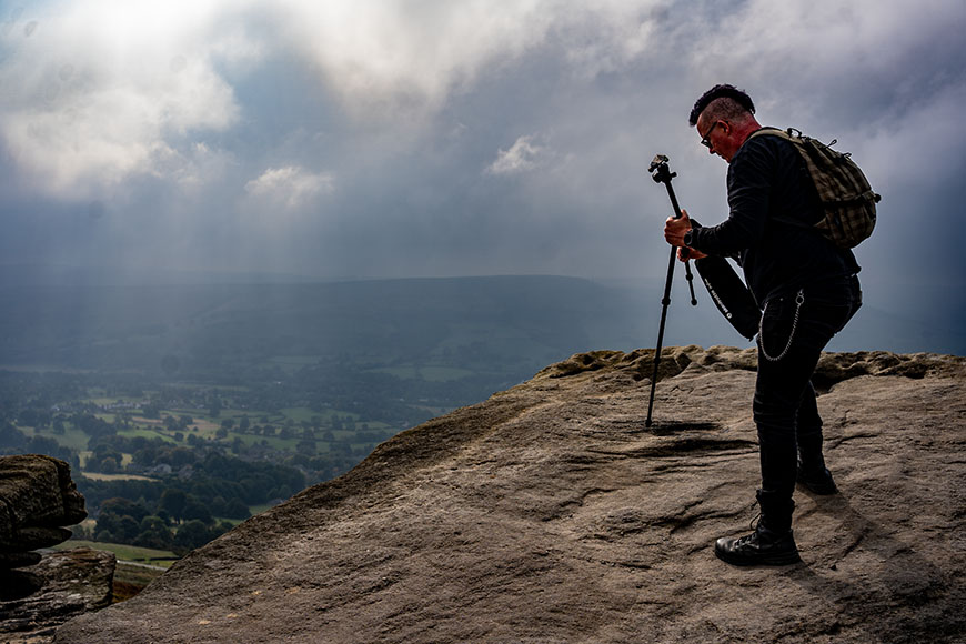 A person with a backpack stands on a rocky cliff, adjusting a camera on a tripod, overlooking a cloudy valley landscape.