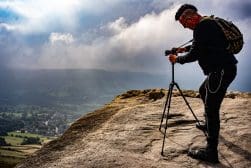 Person with a backpack and camera on a tripod stands on a rocky cliff, photographing a misty landscape below.