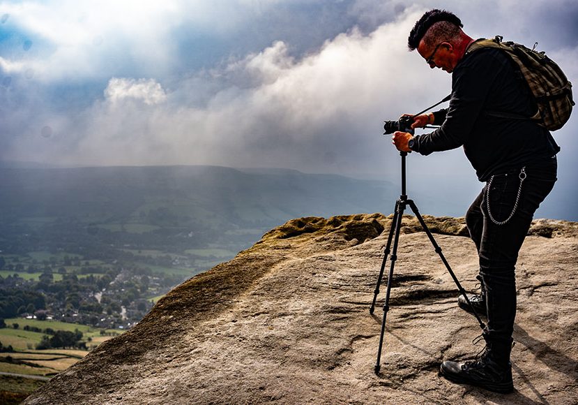 Person with a backpack and camera on a tripod stands on a rocky cliff, photographing a misty landscape below. Person with a backpack and camera on a tripod stands on a rocky cliff, photographing a misty landscape below.