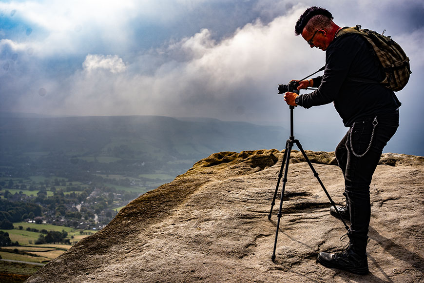 Person with a backpack and camera on a tripod stands on a rocky cliff, photographing a misty landscape below.