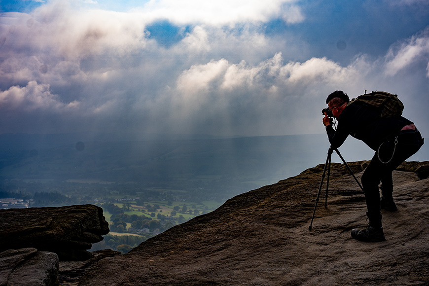Person with a camera on a tripod stands on a rocky ledge, photographing a misty landscape with dramatic clouds and sunlight streaming through.