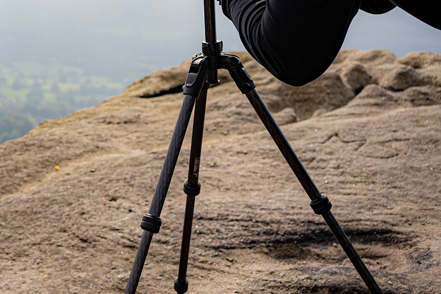 A person adjusts a camera on a tripod set up on a rocky surface, with a hazy landscape in the background.