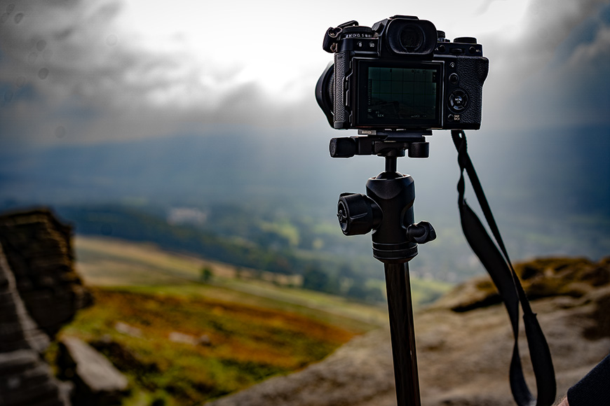 Camera on a tripod overlooking a scenic landscape with hills and cloudy skies in the background.