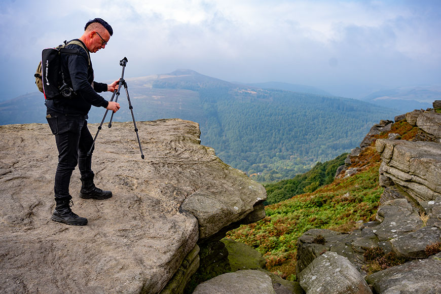 A person sets up a tripod on a rocky cliff with a forested landscape and cloudy sky in the background.