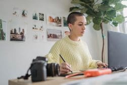 Woman in a yellow sweater working intently on a laptop at a desk with a camera and notebook beside her, in a room decorated with photos.