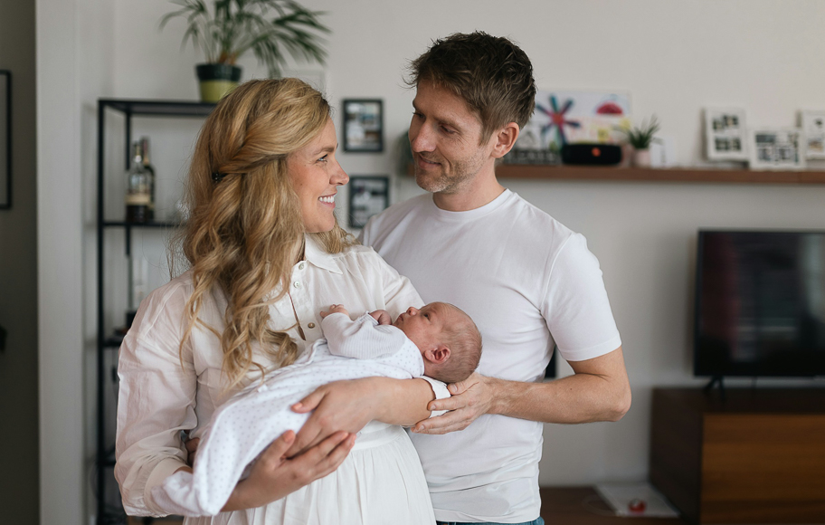 A woman holds a newborn baby while a man stands beside her, both smiling, in a living room with shelves and framed photos in the background.