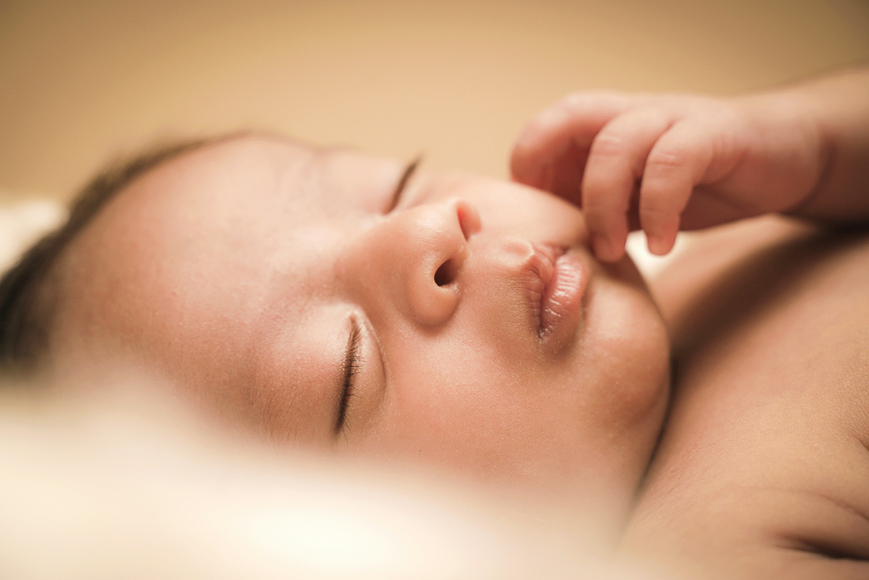 A close-up of a sleeping baby with eyes closed and one hand resting near the face, lying on a soft, light surface.