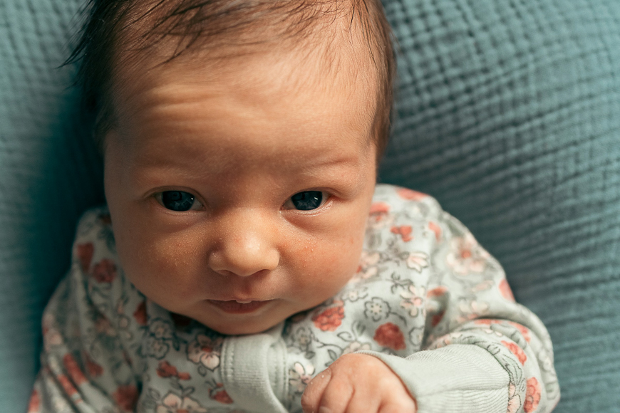 Close-up of a baby with dark hair and brown eyes wearing a floral-patterned outfit, sitting against a textured blue background.