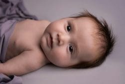 A baby with tousled brown hair lies on a soft surface, partially wrapped in a gray blanket, looking directly at the camera.