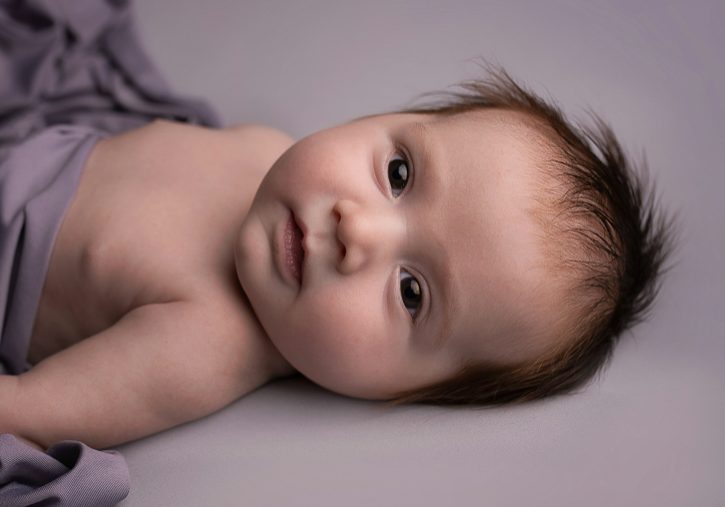 A baby with tousled brown hair lies on a soft surface, partially wrapped in a gray blanket, looking directly at the camera. A baby with tousled brown hair lies on a soft surface, partially wrapped in a gray blanket, looking directly at the camera.