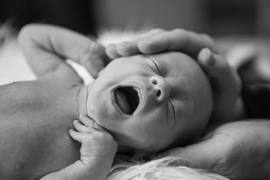 Black and white photo of a newborn baby lying down with mouth open, possibly yawning, while an adult hand gently supports the baby's head.