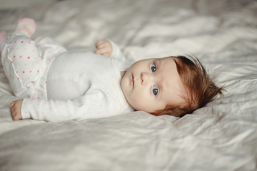 A baby with light skin and brown hair lies on a bed, wearing a white outfit with pink dots, looking towards the camera.