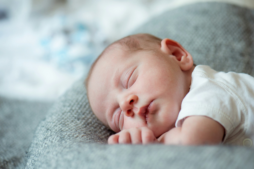 A newborn baby sleeps peacefully on a soft, gray blanket, wearing a white onesie with hands resting near the face.