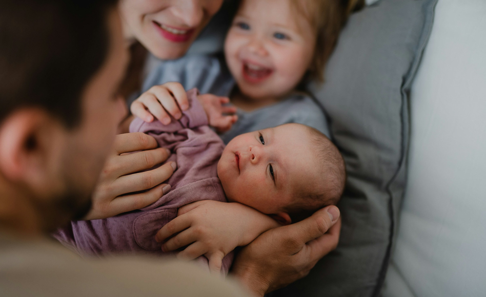 Close-up of a newborn baby being held by an adult, with a smiling woman and young child looking on, all lying on a bed.
