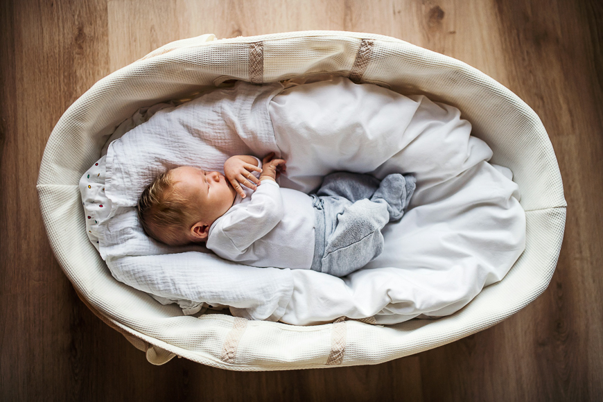 A newborn baby is sleeping on its back in a cushioned crib or bassinet, wearing a white shirt and gray pants, on a wooden floor.