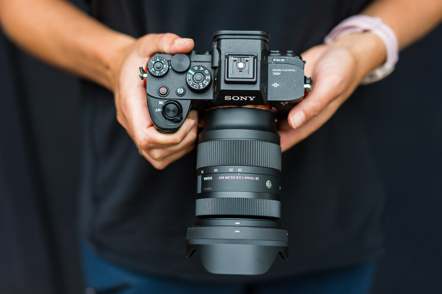 Person holding a Sony digital camera with both hands, showing the top view of the camera and attached lens against a dark background.