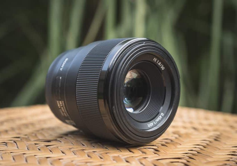 A camera lens labeled "FE 1.8/50" is resting on a woven surface, with a blurred green background behind it.