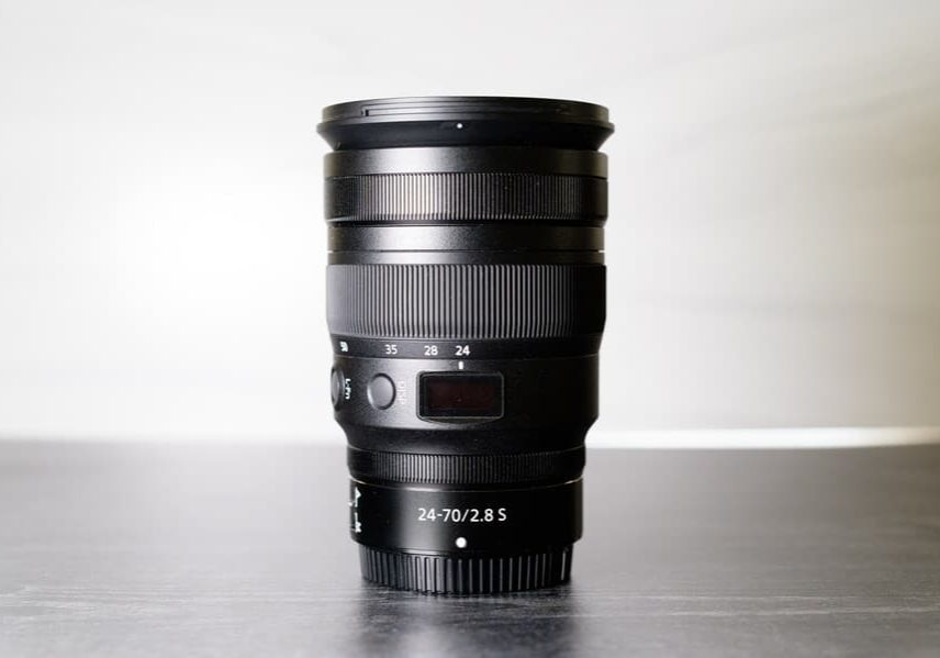 A black camera lens labeled “24-70/2.8 S” stands upright on a gray surface with a neutral background.