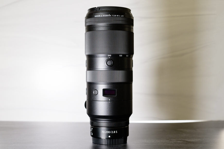 A black Nikon 70-200mm f/2.8 S camera lens stands upright on a reflective surface against a light, blurred background.