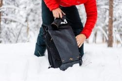 Person in a red jacket placing a camera into a black backpack in a snowy outdoor setting.