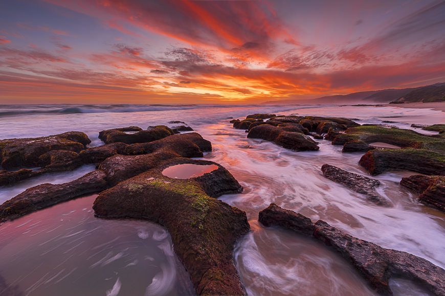 Rocky shoreline with tidal pools at sunset, waves washing over the rocks, and vibrant orange and purple clouds in the sky.