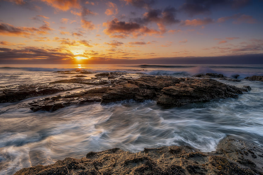 Waves crash against rocky shore under a colorful sunset sky with scattered clouds.