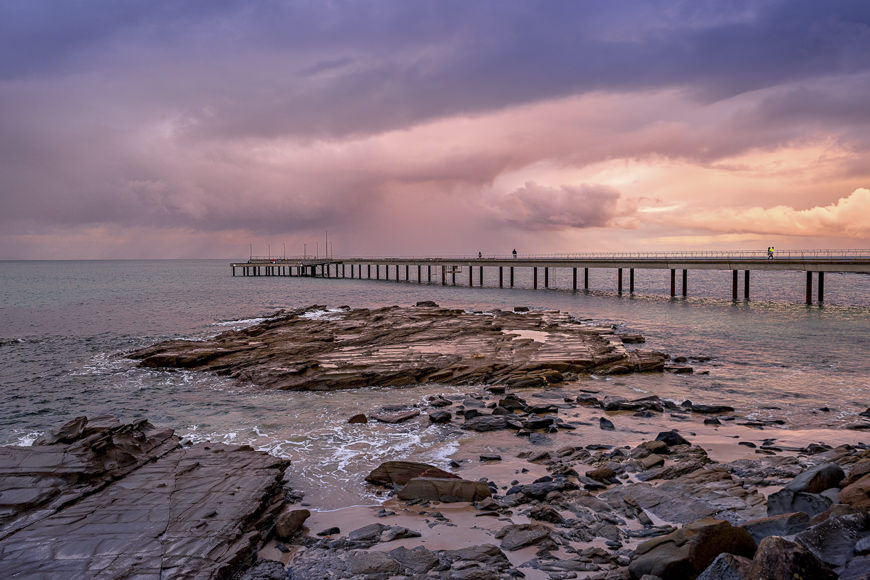 A long pier extends over the sea under a cloudy, sunset sky, with rocky shoreline and gentle waves in the foreground.