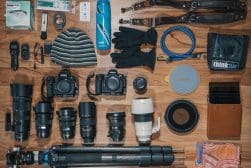 Flat lay of photography gear including cameras, lenses, tripod, gloves, hat, water bottle, camera straps, cleaning supplies, and other accessories arranged on a wooden floor.