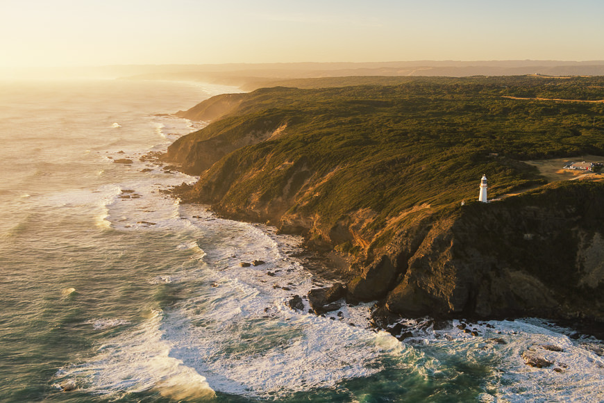 Aerial view of a rugged coastline with waves crashing against cliffs, dense green forest, and a white lighthouse situated near the edge of the land.