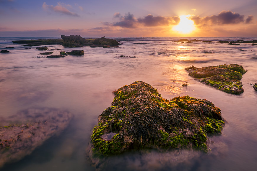 Sunset over the ocean with rocks and seaweed in the shallow water; clouds are scattered in the sky.