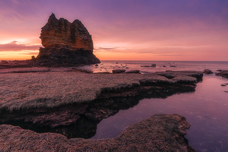 Large rock formation on a coastal shore at sunset, with calm sea, rugged rocks, and purple-orange sky in the background.
