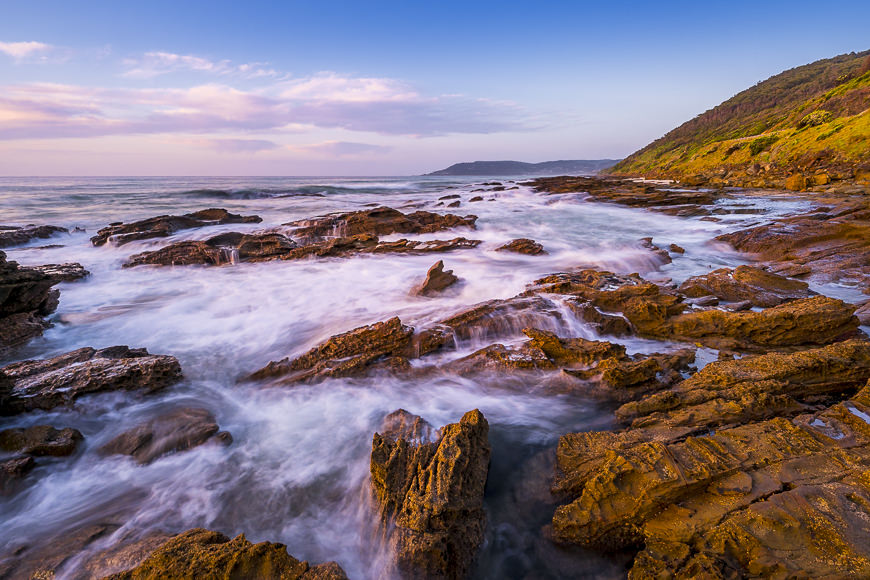 Rocky shoreline with waves crashing against the rocks under a partly cloudy sky, with a green hillside on the right and distant hills along the horizon.