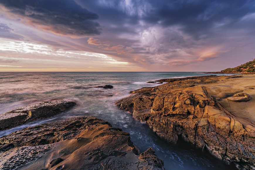 Rocky coastline at sunset with dramatic clouds in the sky and gentle waves washing over the shore.