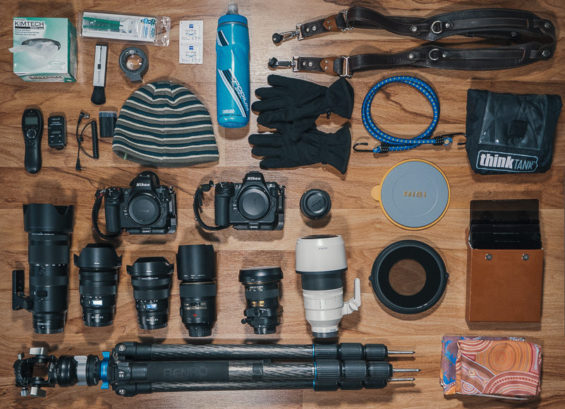 Flat lay of photography gear including cameras, lenses, tripod, gloves, hat, water bottle, camera straps, cleaning supplies, and other accessories arranged on a wooden floor.
