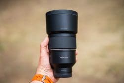 A hand holds a large black camera lens with a wide barrel. The lens is labeled "AF 135 F1.8 FE" and has a matte and textured finish. The person is wearing an orange watch. Background is blurred.