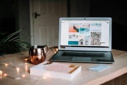 Laptop on a wooden table displaying a mood board, accompanied by a notepad, pen, a mug, and string lights.