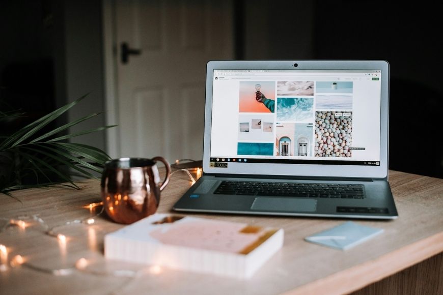 Laptop on a wooden table displaying a mood board, accompanied by a notepad, pen, a mug, and string lights.