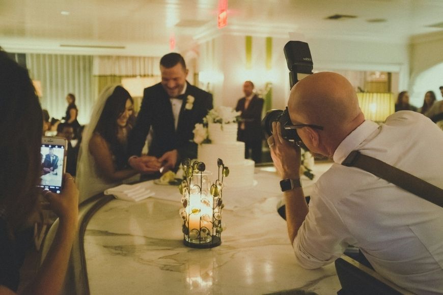 A photographer captures a smiling couple at a wedding reception while a guest takes a photo with a smartphone.