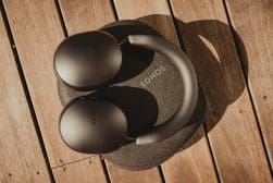 Black Sonos headphones rest on a round Sonos case, placed on wooden floorboards in natural light.