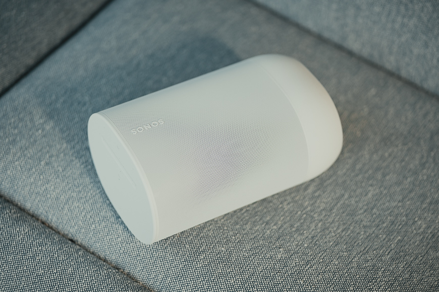 A white Sonos wireless speaker is placed on a light gray upholstered surface.