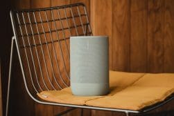 A gray Sonos speaker is placed on a yellow cushion atop a metal wire chair, with a wood-paneled wall in the background.