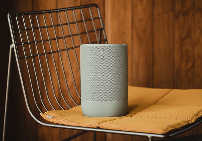 A gray Sonos speaker is placed on a yellow cushion atop a metal wire chair, with a wood-paneled wall in the background. A gray Sonos speaker is placed on a yellow cushion atop a metal wire chair, with a wood-paneled wall in the background.